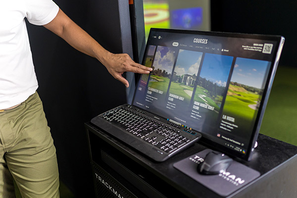 A man gestures towards a computer screen showing a golf game, illustrating the use of simulators in golf training.