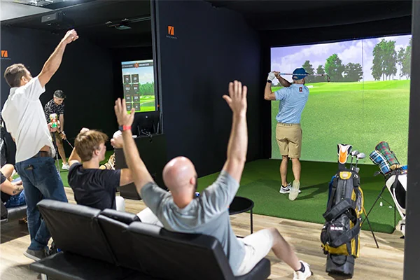 Group of individuals playing golf in a simulator room, featuring a TV screen showcasing a virtual golf course.