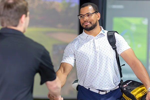 Two men shake hands on a golf course, representing camaraderie and collaboration in indoor golf practice discussions.