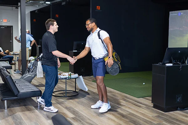 Two men shaking hands in a golf room, engaged in a conversation about effective indoor golf practice strategies.
