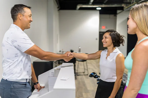 A man and woman greet each other with a handshake at a reception desk, related to indoor golf in San Antonio.