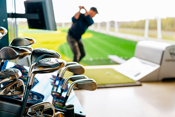 A man practices his golf swing indoors, surrounded by numerous golf clubs, highlighting a golf simulator setting.