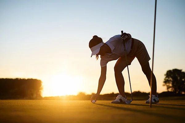 A woman leans down to set her golf ball on the green, highlighting the accuracy of golf simulator scores versus real play.