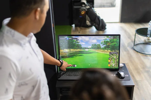 A man in a white shirt points at a golf simulation on a monitor, inside a room with golf equipment in the background. The atmosphere is focused.
