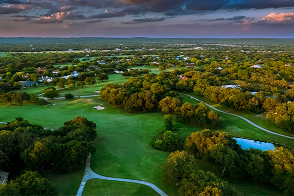 Aerial view of a vast, lush green landscape dotted with trees and a winding path, under a dramatic, cloudy sunset sky. The scene conveys a peaceful, serene atmosphere.