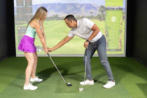 A man and woman play golf on a simulator, with the screen showing a virtual golf course as they prepare to take their shots.