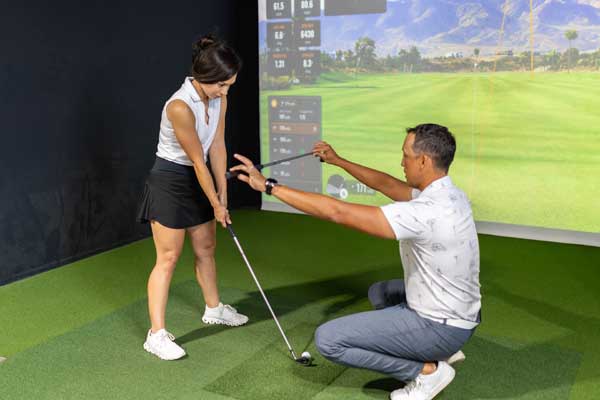  A man and woman play golf on a simulator, with the screen showing a virtual golf course as they prepare to take their shots.