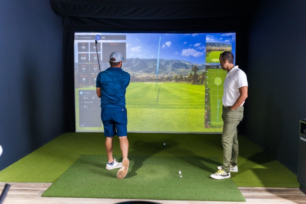 Two men engaged in a golf simulation game inside a room, focusing on their swings and the virtual screen ahead.