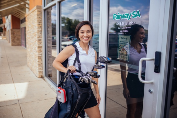 Outside a golf business, a woman carries a golf bag, preparing for her indoor golfing session.