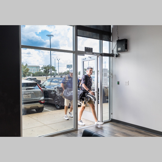 A person carrying golf clubs enters a modern building through a glass door, with cars visible outside.