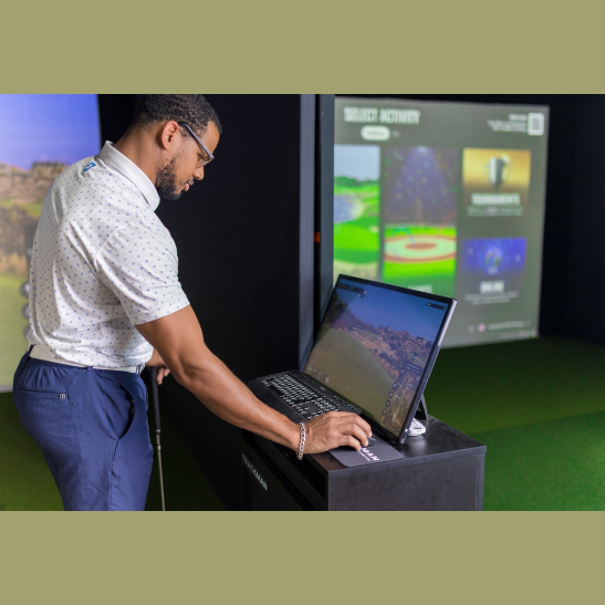 A man adjusts settings on a computer while preparing to play golf in a virtual simulator.