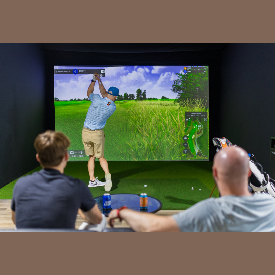 A golfer swings a club in a virtual golf simulator while two spectators watch from a nearby seating area.