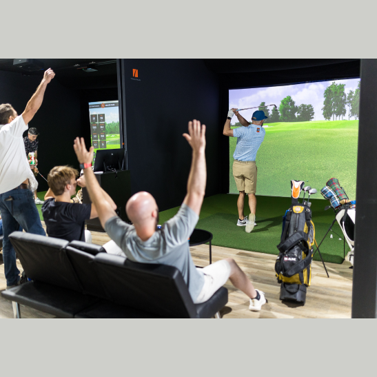A golfer swings at a virtual golf simulator while spectators cheer from a nearby couch.