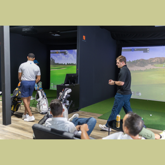 A golfer prepares to swing in a virtual golf simulator while others relax on a couch nearby.