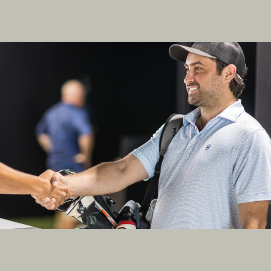 A man in a light blue polo shirt shakes hands with another person, holding a golf bag over his shoulder.