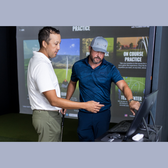 Two men discuss golf practice techniques while looking at a computer screen in a simulated golf environment.