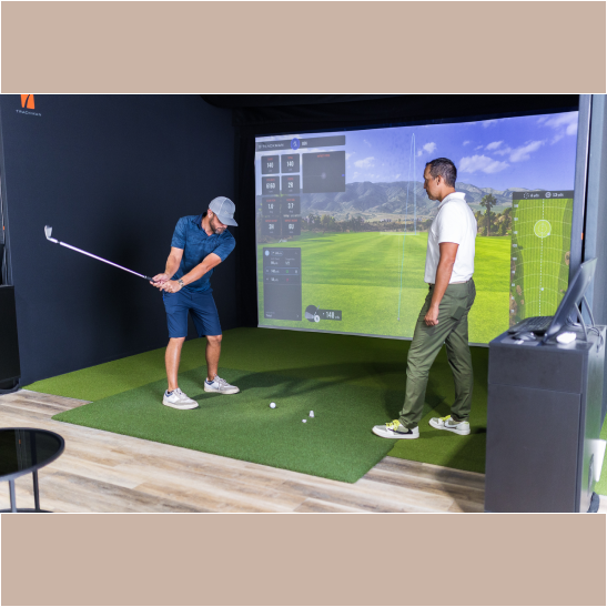 A golfer swings a club while an instructor observes in a virtual golf simulation setup.