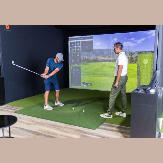 A golfer swings a club while an instructor observes in a virtual golf simulation setup.