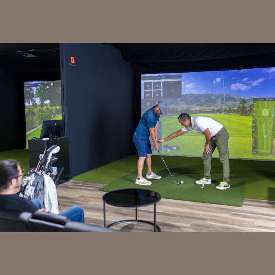 A golfer receives instruction on their swing technique in a virtual golf simulator setting, with a coach demonstrating adjustments.
