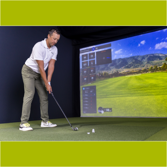 A golfer prepares to swing in a virtual golf simulator, with a large screen displaying a scenic course in the background.