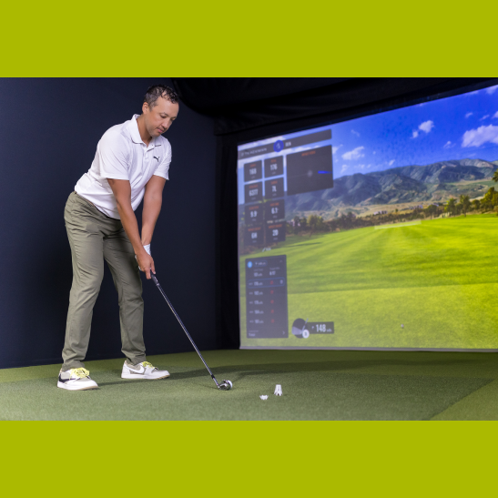 A golfer prepares to swing in a virtual golf simulator, with a large screen displaying a scenic course in the background.